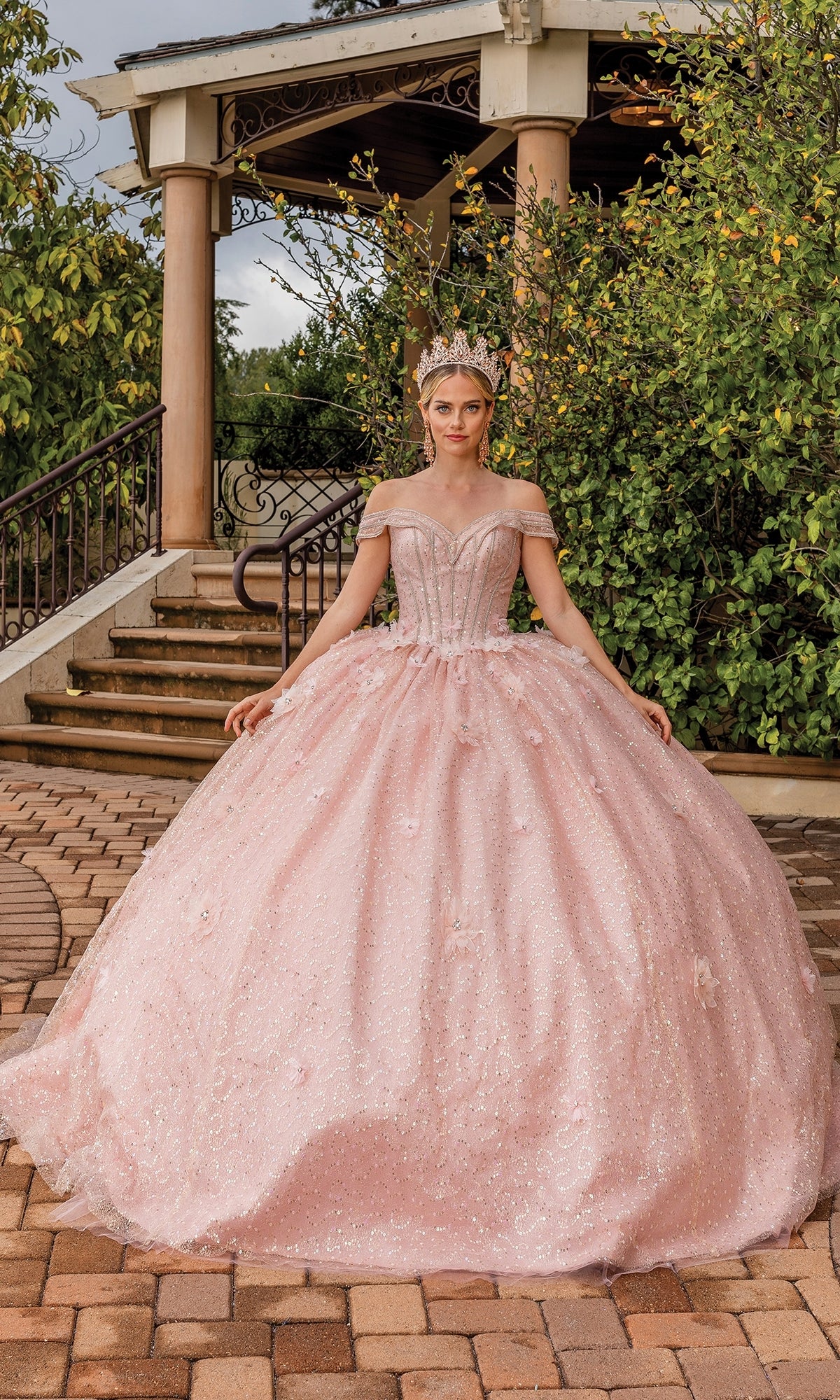 A woman wearing the Quinceanera Dress 1868 by Dancing Queen—pink, off-the-shoulder with a full skirt—stands outdoors on a stone path in front of a gazebo, crowned and radiant.